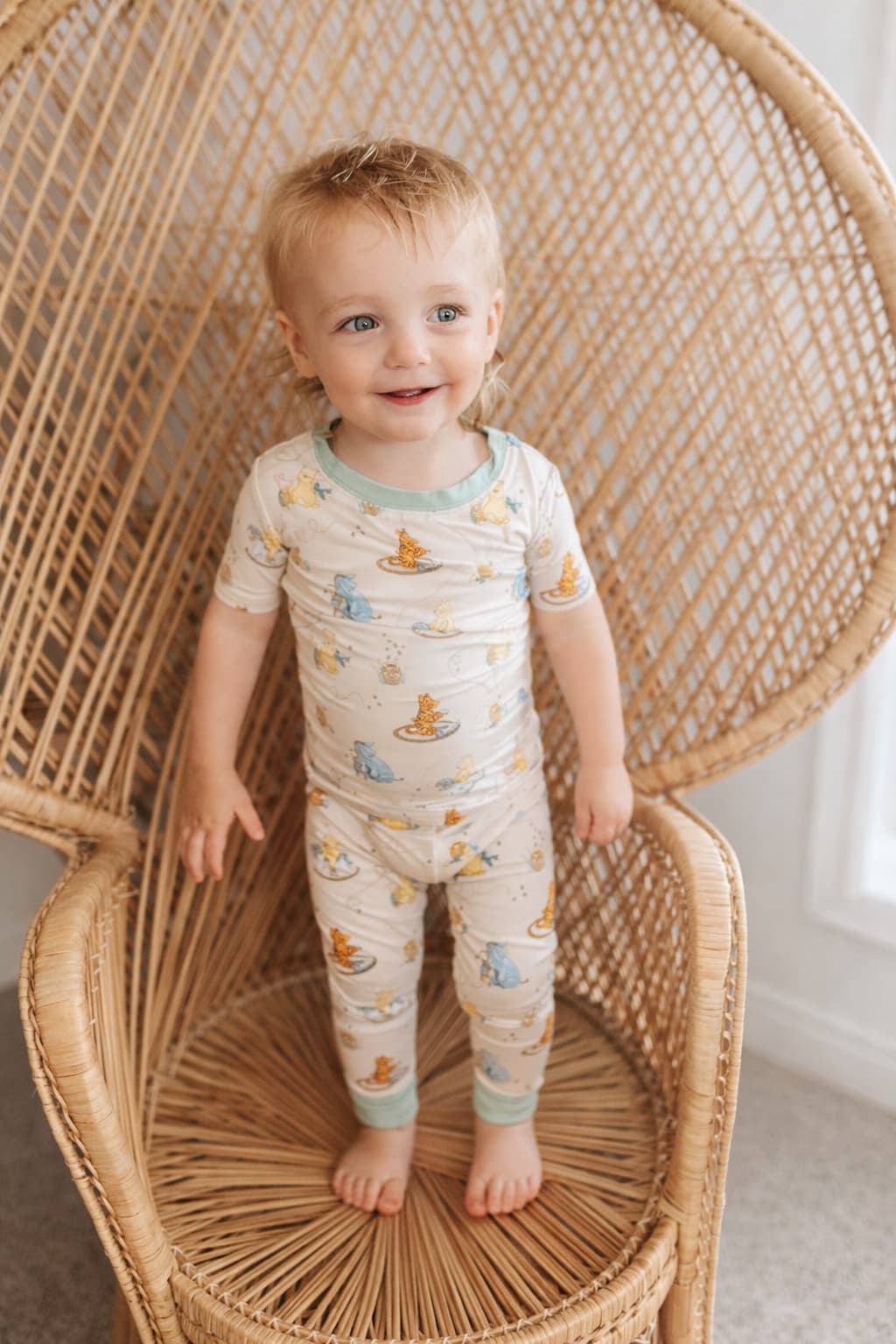 Child wearing bamboo pajamas with classic pooh, eeyore, tigger, and piglet print standing on a wicker chair.